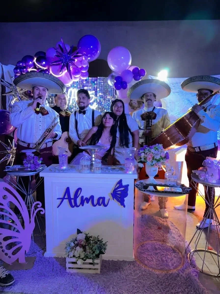 Mariachis con trompeta y guitarrón junto a una quinceañera y su familia, frente a una mesa con globos violetas y plateados